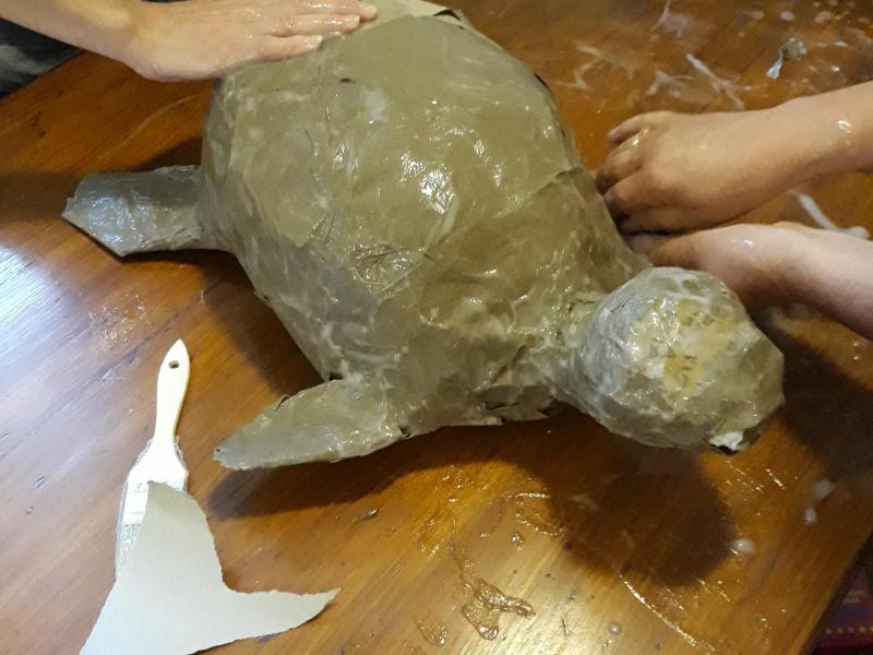 Postać żółwia lepiona z moczonego papieru przez dwie kobiety na drewnianym stole. W tle miska z materiałem.