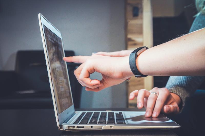Ręka jednaj osoby wskazująca palcem na coś na ekranie laptopa. Dłoń innej osoby dotyka gładzika.